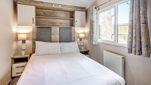 A bedroom with a bed, nightstands, a headboard, a window with curtains, and a radiator. - Silver Caravan with parking (Great Yarmouth)