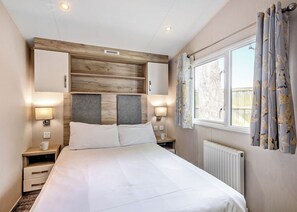 A bedroom with a bed, nightstands, a headboard, a window with curtains, and a radiator. - Silver Caravan with parking (Great Yarmouth)