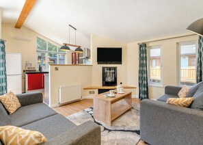 A modern living room with grey sofas, a wooden coffee table, a flat-screen TV, and a kitchen area with red cabinets and a white refrigerator.