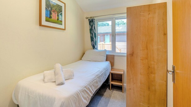 Una cama individual con ropa de cama blanca, una mesita de noche pequeña, una ventana con cortinas y una puerta.