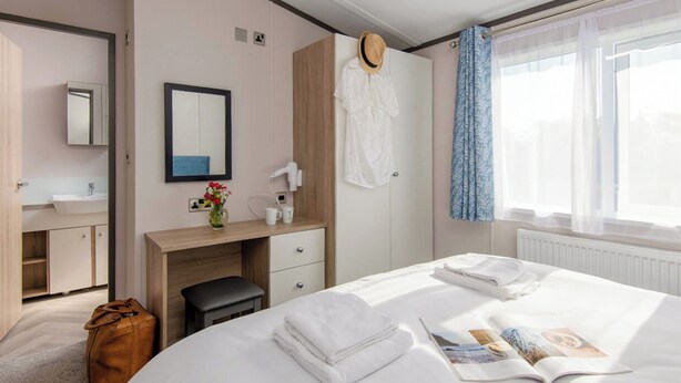 A hotel room with a bed, a desk, a mirror, a vanity with a sink, a suitcase, and a window with curtains. - Deluxe 4, Slps 8, parking, ensuite, decking (Near Weymouth)