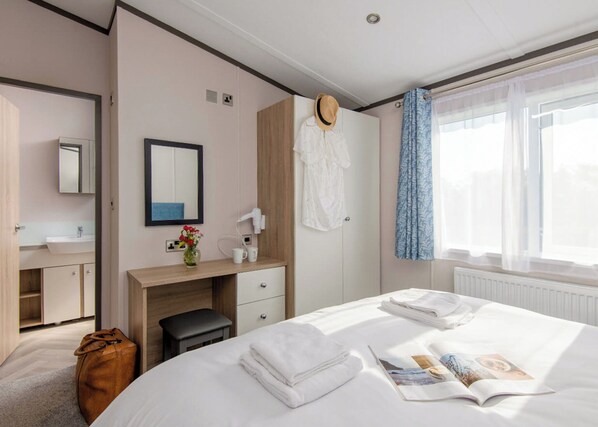 Une chambre d’hôtel avec un lit, un bureau, un miroir, une coiffeuse avec un lavabo, une valise et une fenêtre avec des rideaux.