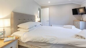 Quarto de hotel com cama, mesa de cabeceira, luminária, televisão e cômoda.