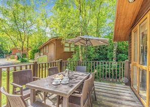 Una terraza de madera con muebles de mimbre y una mesa preparada para el té, rodeada de abundante vegetación y una cabaña de madera.