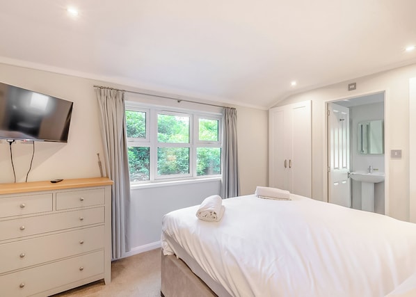Un dormitorio con cama, televisor, cómoda, ventana con cortinas y puerta que da al baño.