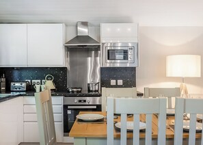 A modern kitchen with white cabinets, a stainless steel range hood, an oven, a microwave, and a dining area with a table and chairs.