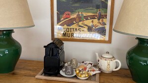 A framed poster of a mountainous landscape with the text "MUJOTAS - MURAGU" on a wooden table with two green lamps, a coffee maker, teapots, and a ceramic mug.