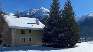 Una casa cubierta de nieve con una chimenea, un gran abeto y montañas al fondo.