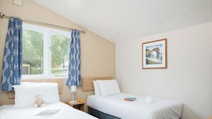 Ein Schlafzimmer mit zwei Betten, einem Fenster mit Vorhängen, einem Nachttisch mit Lampe und einem gerahmten Bild an der Wand.