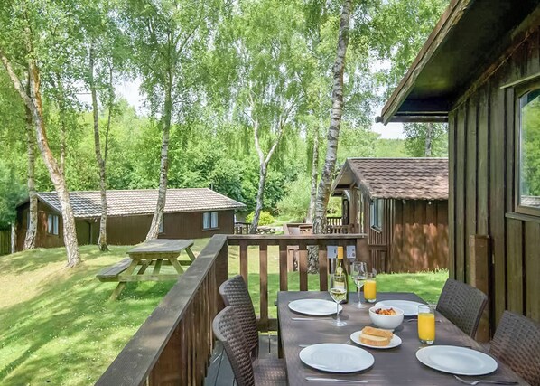 A wooden deck with a table set for breakfast, surrounded by trees and a small building in the background.