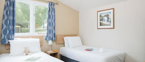 A bedroom with two beds, a window with curtains, a bedside table with a lamp, and a framed picture on the wall.