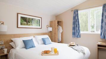A bedroom with a bed, a bedside table with a cup and a tray of croissants and juice, a wardrobe, a window with curtains, and a framed picture on the wall.