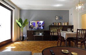 Ein Wohnzimmer mit Fernseher, Esstisch, Stühlen, einem Bücherregal und einer Zimmerpflanze.