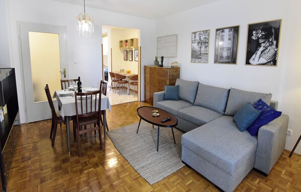 Ein Wohnzimmer mit grauem Sofa, Esstisch aus Holz und Stühlen. In der Mitte steht ein Couchtisch.