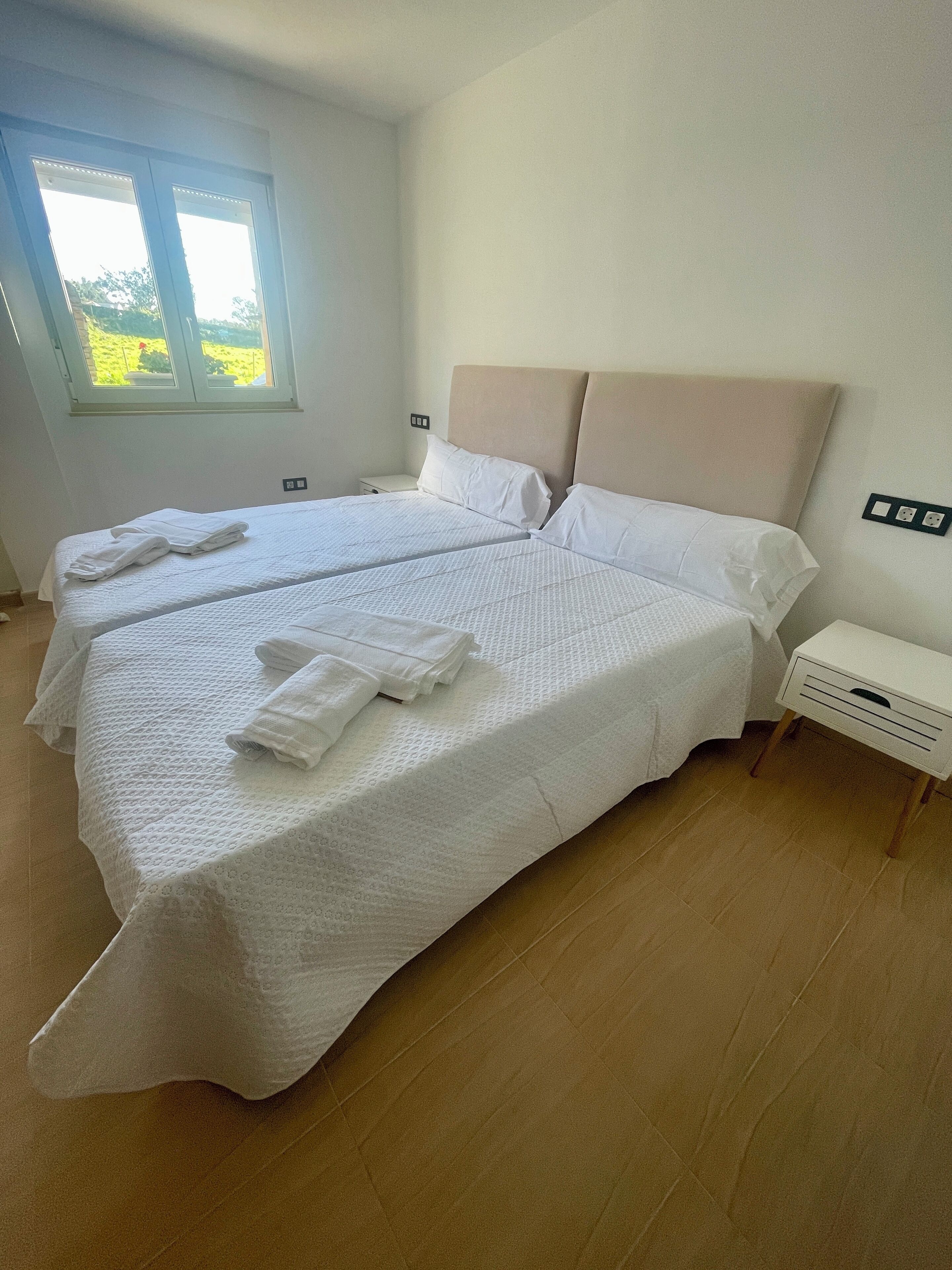 Un lit double avec une literie blanche et une tête de lit, une table de chevet avec une lampe, et une fenêtre donnant sur un paysage verdoyant.