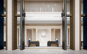 Uma área de recepção de hotel com paredes de mármore, um balcão de atendimento e uma placa com a inscrição 'VOCO AN IHG HOTEL'.