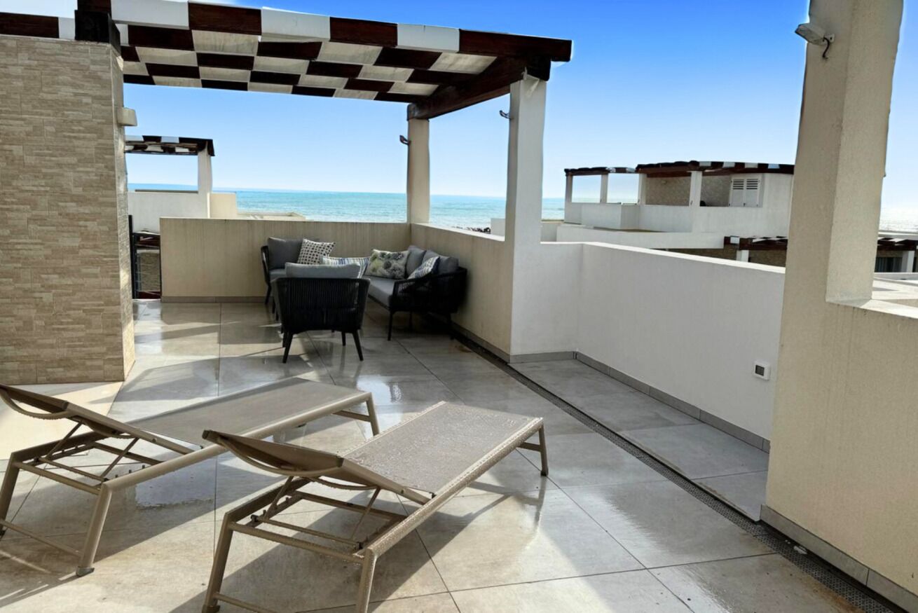 A rooftop terrace with a checkered ceiling, a seating area with a sofa and chairs, and a view of the ocean.