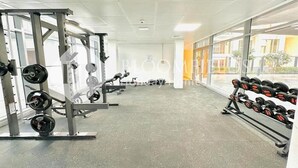 A gym with weightlifting equipment, large windows, and a view of buildings outside. - Bloomfields Pretty Stu in Oasis  (Abu Dhabi)