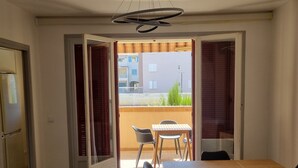 Een open schuifdeur leidt naar een balkon met een tafel en stoelen, een plafondlamp en een koelkast in de aangrenzende kamer.