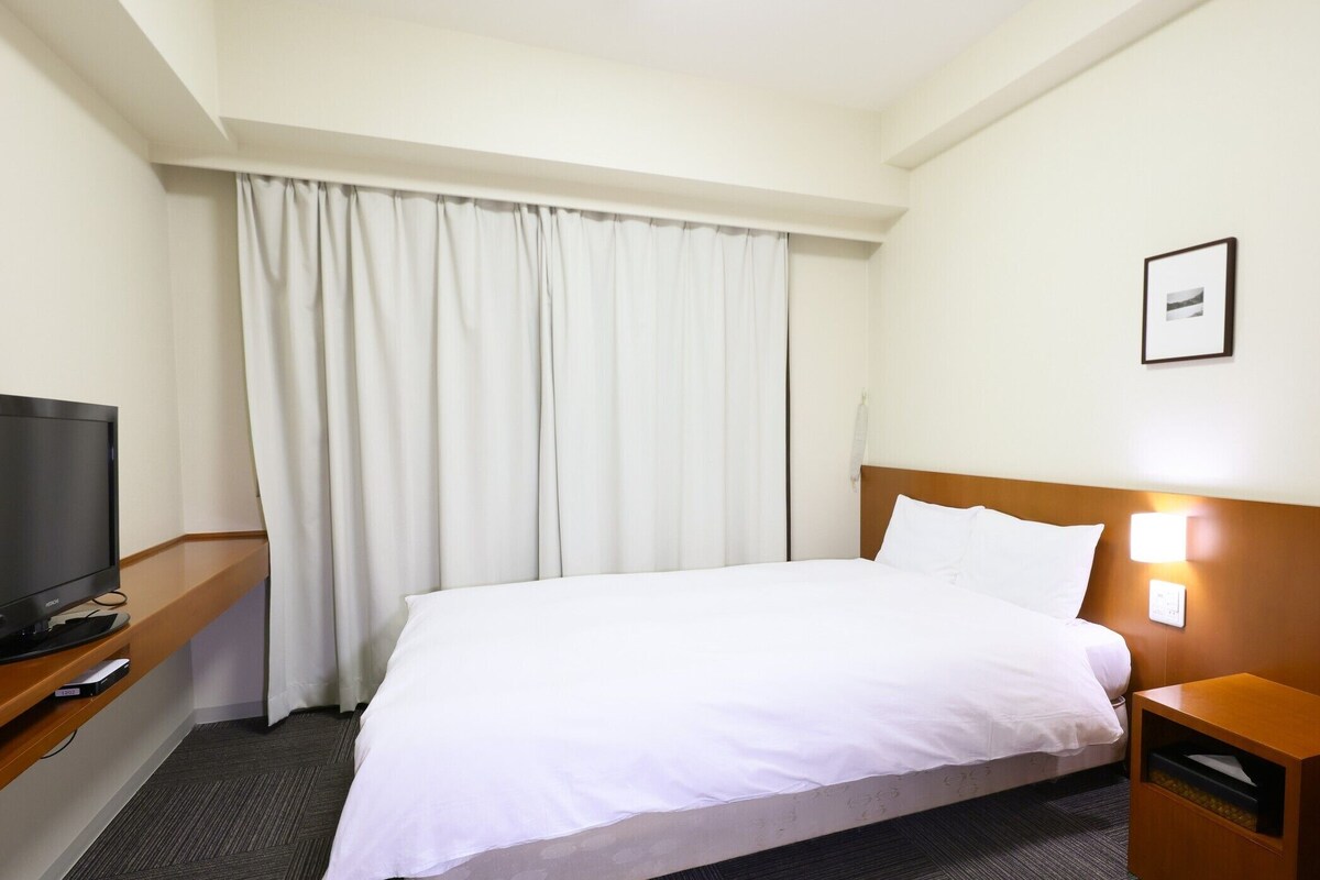 A hotel room with a bed, a TV, a bedside table, a lamp, and a picture on the wall.