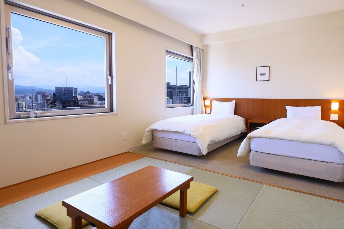 A hotel room with two beds, a wooden table, tatami mats, and a city view.