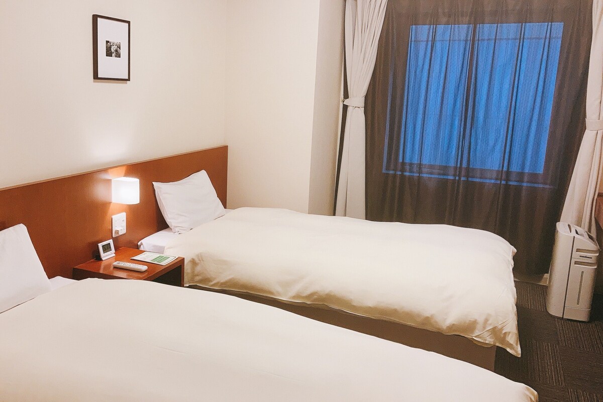 A hotel room with two beds, a nightstand with a lamp, a clock, and a window with curtains.