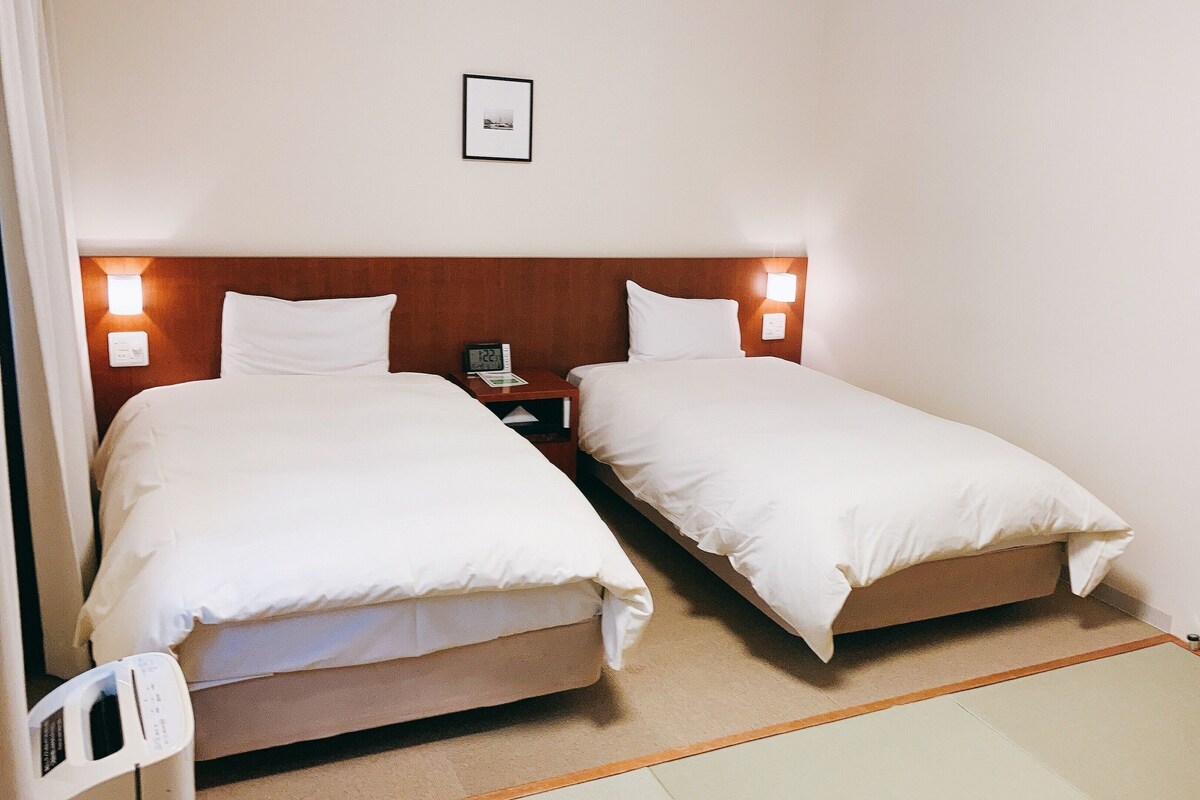 A hotel room with two single beds, a bedside table with a clock, and a wall-mounted picture.