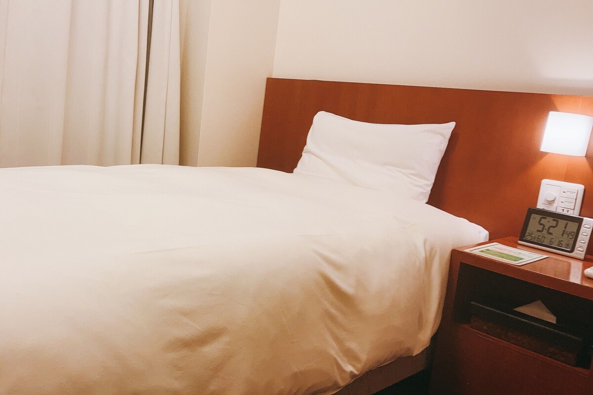 A hotel room with a bed, a nightstand with a clock, and a wall-mounted light.