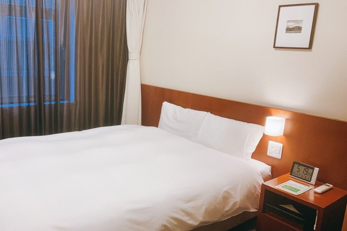Una habitación de hotel con cama, mesita de noche con un reloj y un control remoto, y una ventana con cortinas.