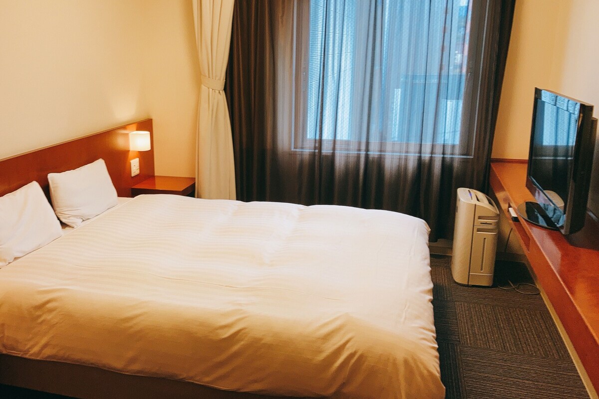 A hotel room with a large bed, a bedside table with a lamp, a TV, and a window with curtains.