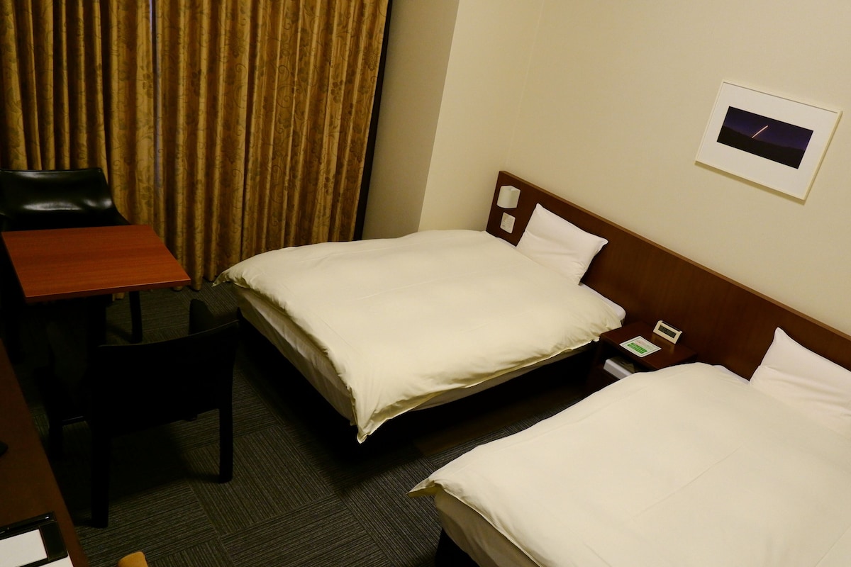 A hotel room with two beds, a desk, a chair, and a nightstand with a clock.