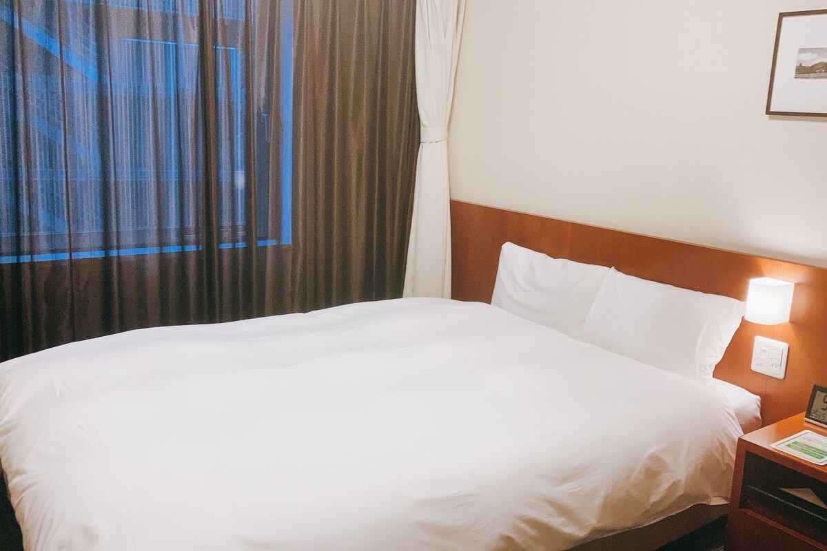 A hotel room with a bed, a nightstand with a clock, and a wall-mounted picture.