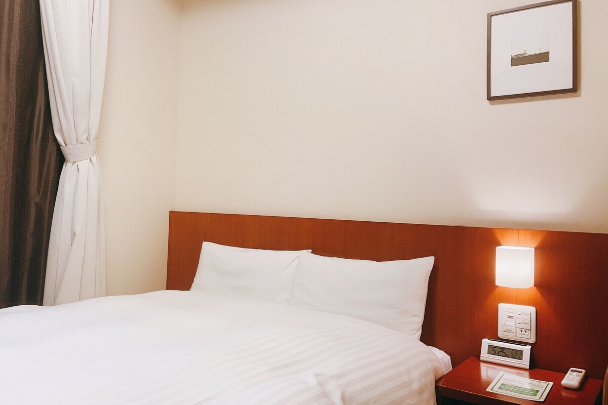 A hotel room with a bed, a nightstand with a lamp, a framed picture on the wall, and a window with curtains.