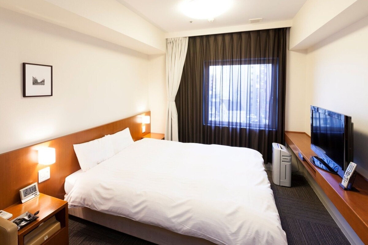 A hotel room with a bed, bedside lamps, a TV, a desk with a computer, and a window with curtains.