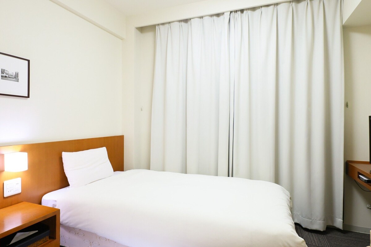 A hotel room with a bed, a nightstand, a TV, and a curtain.
