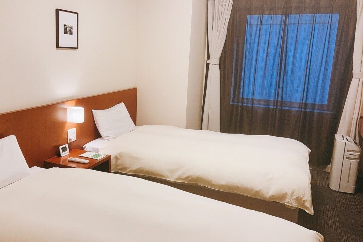 A hotel room with two beds, a bedside table with a lamp, a framed picture on the wall, and a window with curtains.