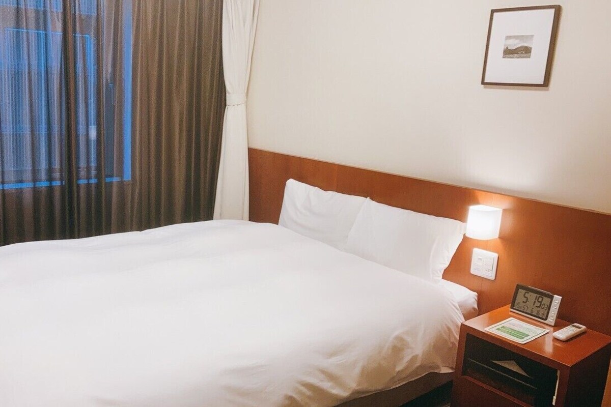 A hotel room with a bed, a nightstand with a clock and a remote, and a window with curtains.