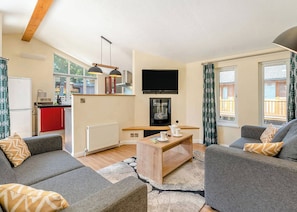 A modern living room with grey sofas, a wooden coffee table, a flat-screen TV, and a kitchen area with red cabinets and a white refrigerator.