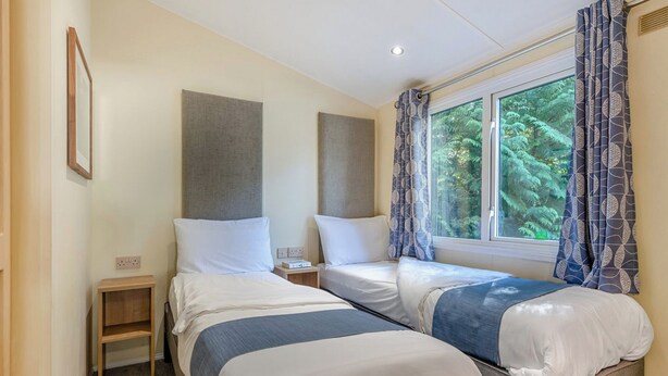 A twin room with two beds, a small wooden nightstand, a window with curtains, and a framed picture on the wall. - Superior Lodge with ensuite & decking (Near Weymouth)