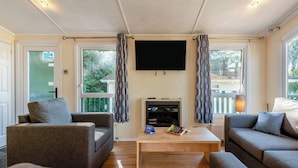 A living room with two sofas, a coffee table, a flat-screen TV, and large windows with curtains. - Superior Lodge with ensuite & decking (Near Weymouth)
