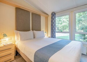 A hotel room with a bed, a nightstand, a lamp, and a window with a view of greenery. - Superior Lodge with ensuite & decking (Near Weymouth)