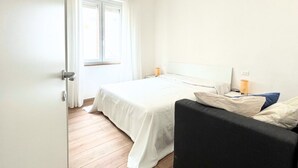 Un dormitorio con cama, mesita de noche, lámpara, sofá y ventana.
