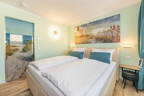 A bedroom with a bed, a painting of a beach scene, a wall-mounted light, and a small table with a chair. - Villa Kamadori 07: Stylish apartment with balcony, 150 m to the beach (Ahlbeck (Seebad))