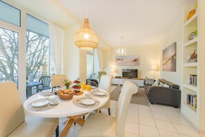 A modern living room with a dining area, featuring a round table set for breakfast, a sofa, and a bookshelf. - Südwind 13—two bedrooms in a prime location, sauna, pool, and balcony (Zempin)