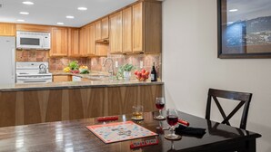 Una cocina con armarios de madera, un microondas y un lavabo. Hay una mesa con un juego de mesa, vasos y una botella.