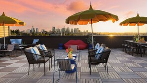 Eine Dachterrasse mit Sitzgelegenheiten im Freien, Sonnenschirmen und Blick auf eine Stadtlandschaft im Sonnenuntergang.