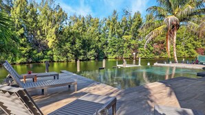 A wooden deck with lounge chairs and a table, overlooking a clear pool surrounded by lush greenery and a palm tree. - Waterfront, Infinity Pool, Dock, Outdoor Bar, Beach Access | Riverside Veranda (Fort Lauderdale)