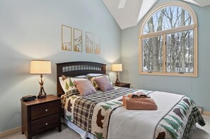 Een slaapkamer met een groot bed, twee nachtkastjes, een raam met uitzicht op bomen, en drie kunstwerken aan de muur.