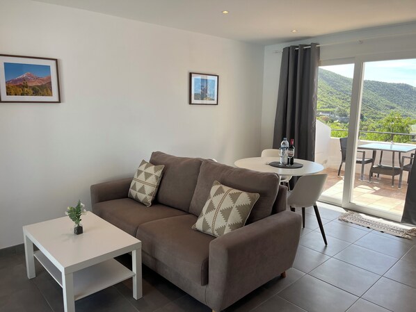 A modern living room with a brown sofa, a white coffee table, and a dining area with a table and chairs. There are two framed pictures on the wall, and a view of a mountainous landscape through a large window. - Ventoso D Apartment with sea view in the nature at 300m altitude 3km from the beach (Santiago del Teide)