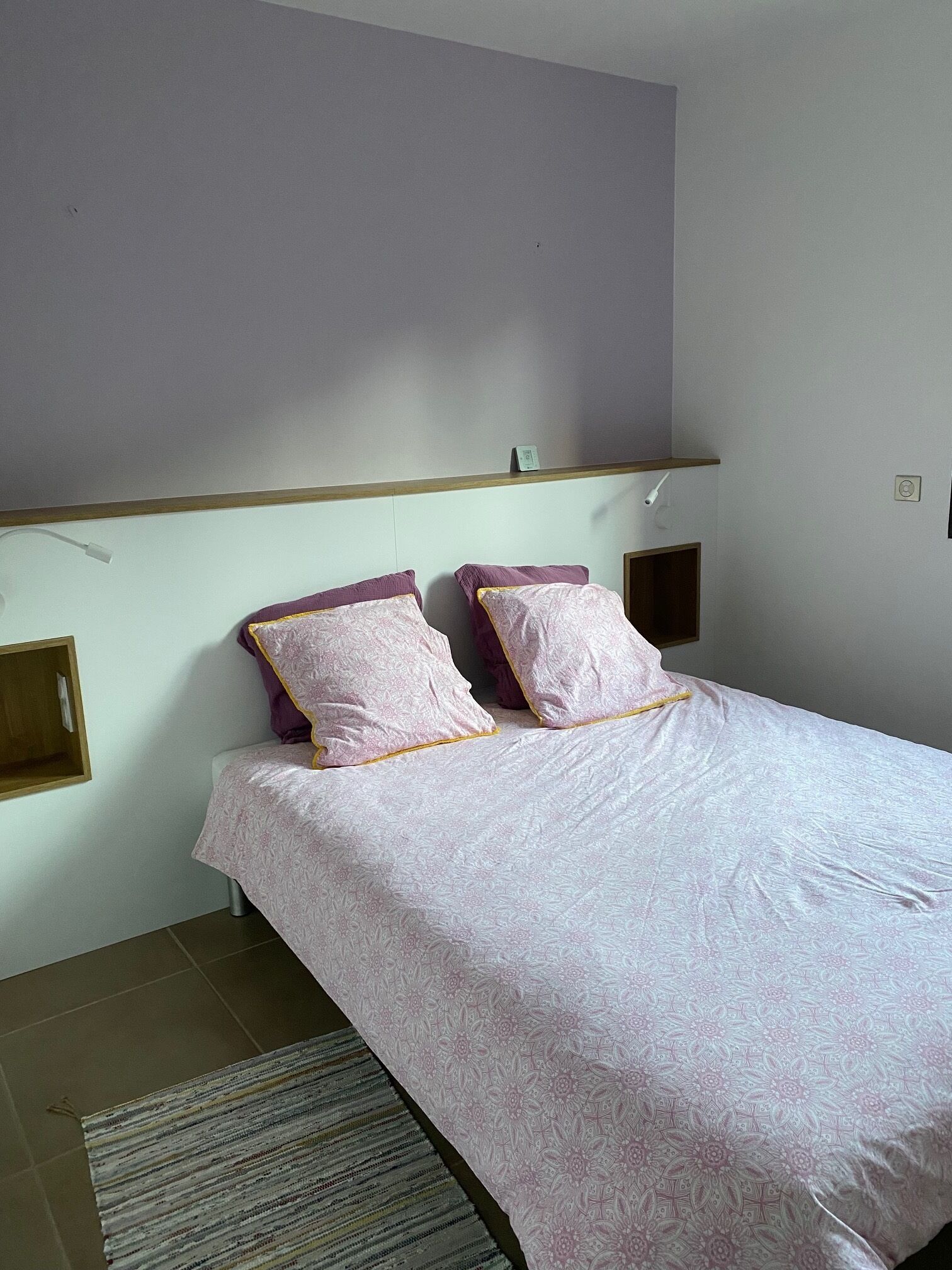 Ein Einzelbett mit einem gemusterten Bettbezug und zwei Kissen, ein hölzernes Kopfteil, eine kleine Ablagefläche und eine wandmontierte Lampe.